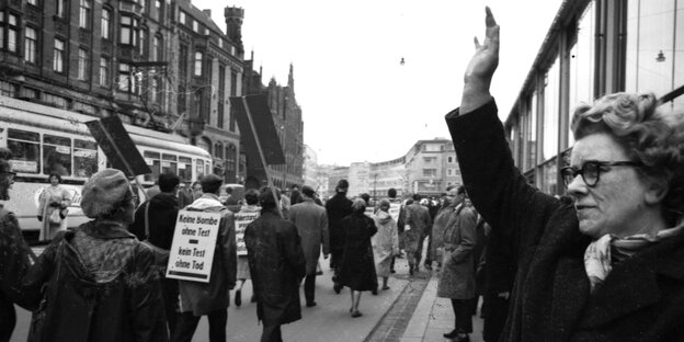 Schwarz-weiß-Foto, eine Frau steht mit erhobener Hand am Rande einer Demonstration. Die Teilnehmer wurden von hinten fotografiert - einer trägt ein Schild auf dem Rücken: Keine Bombe ohne Test - kein Test ohne Tod
