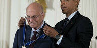 Barack Obama und Daniel Kaheman tragen schwarze Anzüge. Obama legt Kahneman von hinten eine Medaille um den Hals.